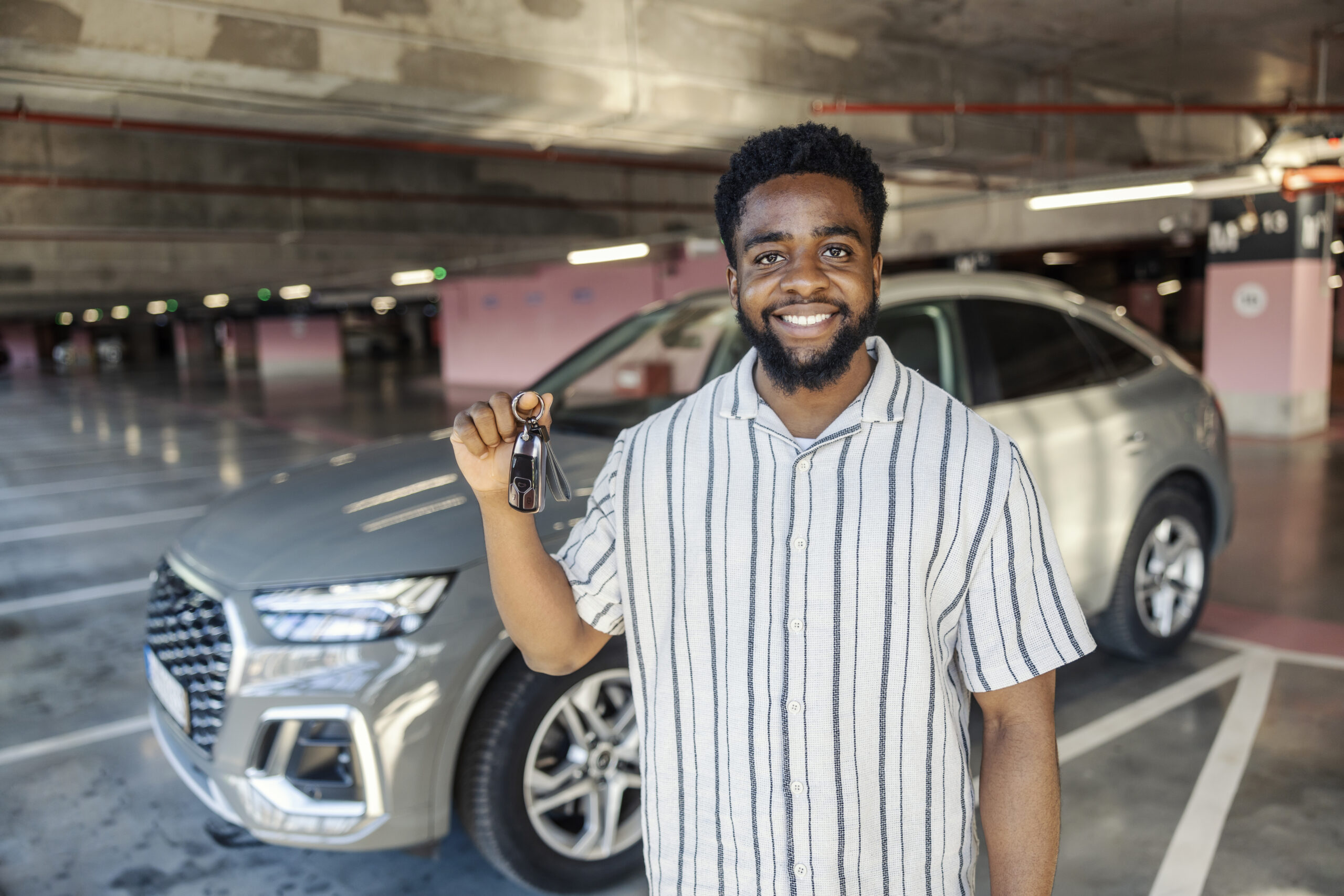 portrait of a happy young man standing at public garage with car keys with his classy and new car in a blurry background.